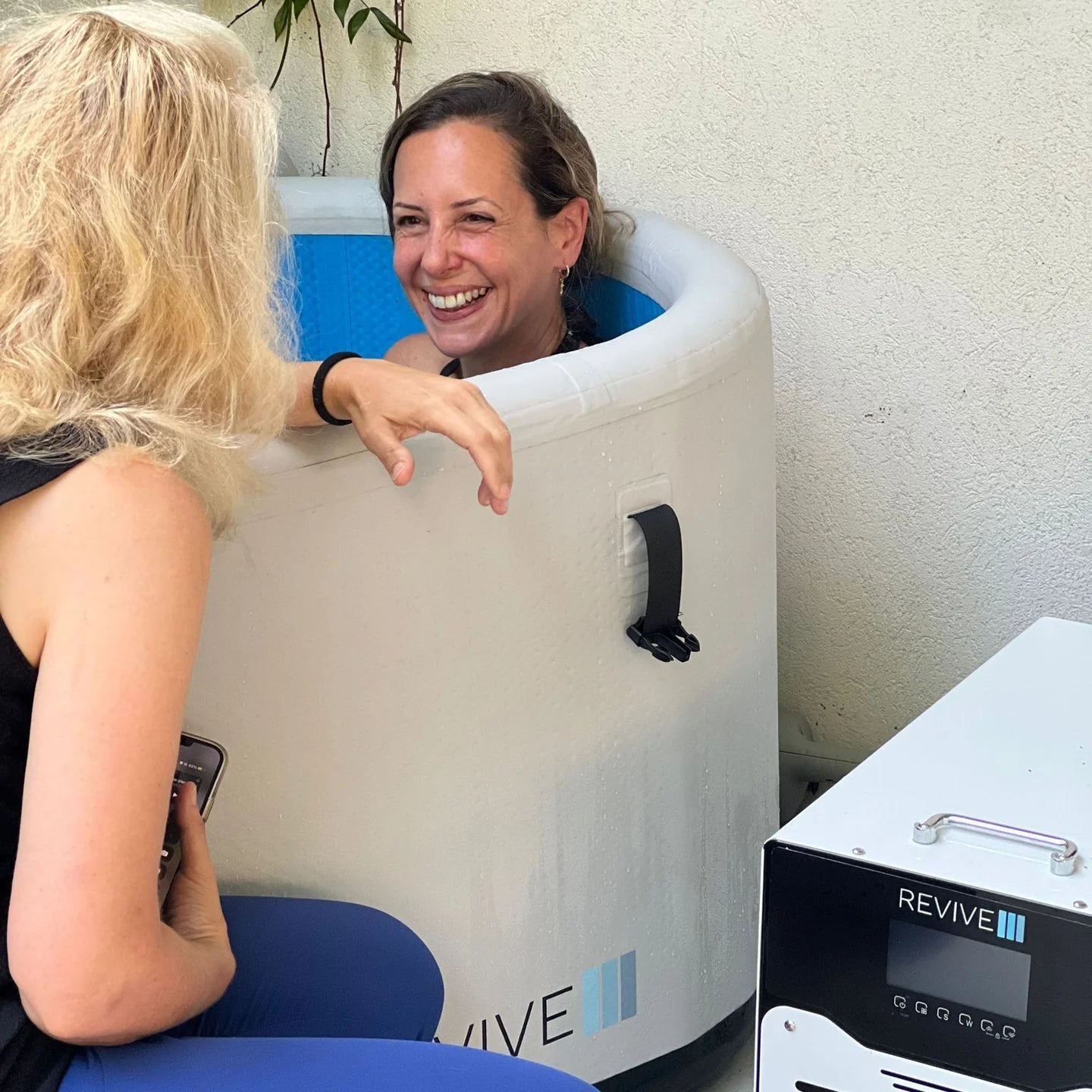 Two women interacting with a 'REVIVE' cryosauna chamber.