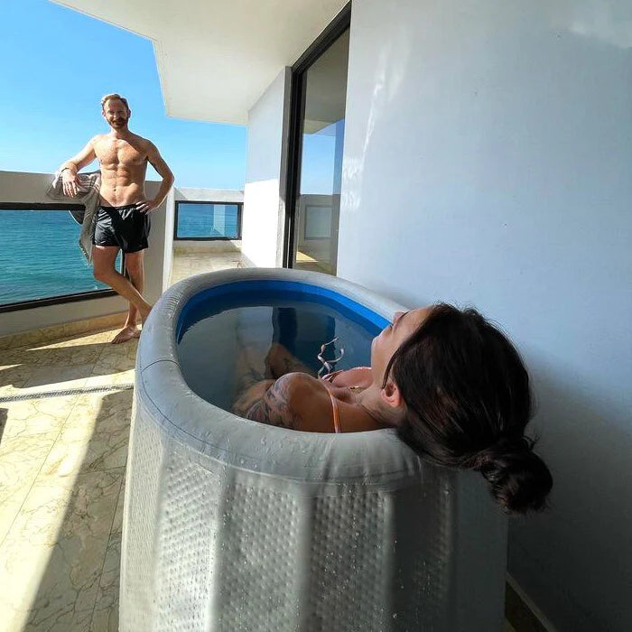 Woman relaxing in a hot tub with a man standing nearby on a balcony overlooking water.