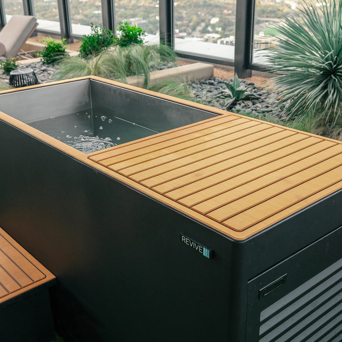 Outdoor hot tub with wooden deck and 'REVIVE' branding, surrounded by plants and a cityscape.