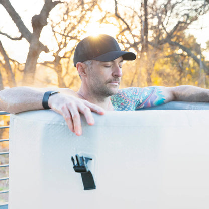 Man with tattoos leaning on a white object outdoors with trees in the background