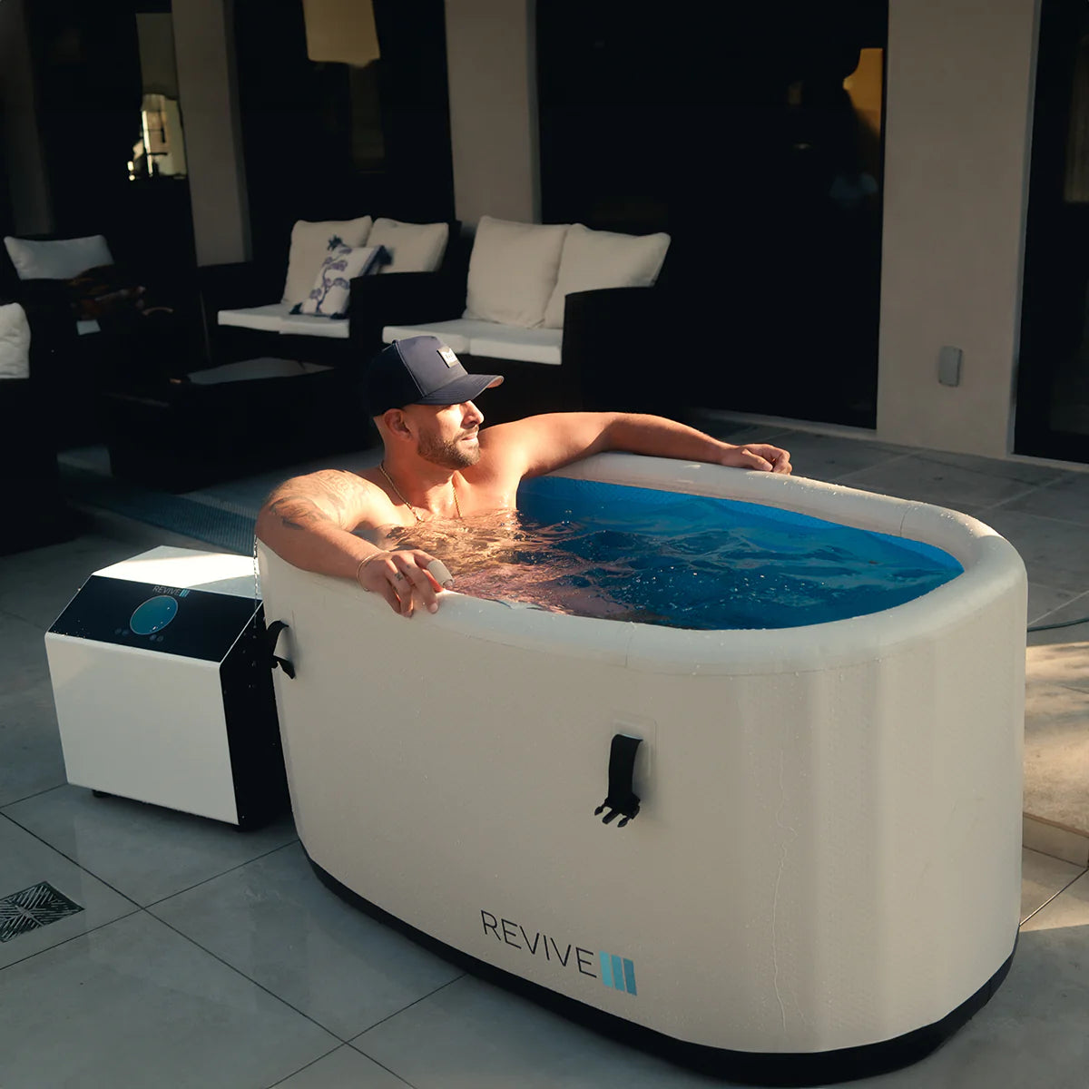 Man relaxing in a white inflatable cold tub labeled 'REVIVE' in a modern living room.