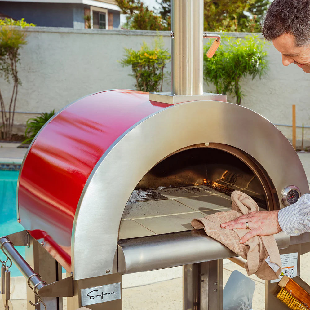 Person using a red and silver outdoor pizza oven with a pool and garden in the background.
