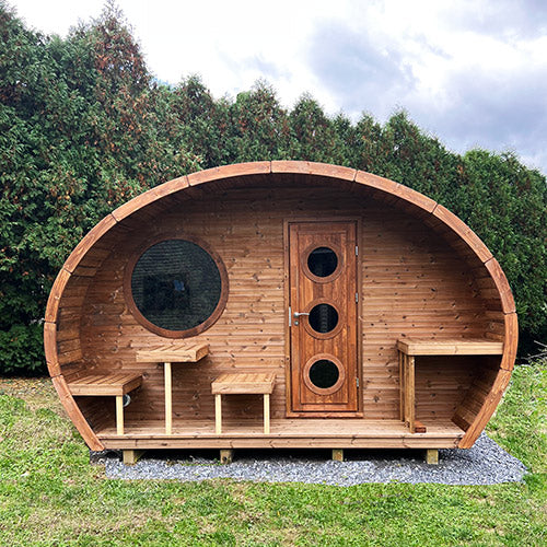 Wooden barrel-shaped cabin with a door and windows on a grassy area with trees in the background.