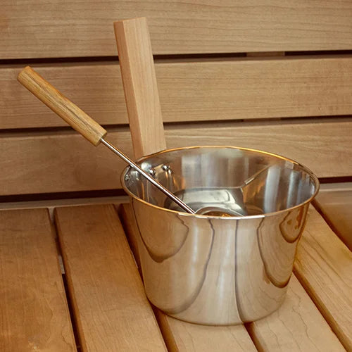 A stainless steel sauna bucket with a wooden handle and a wooden scoop resting inside, set on a wooden slatted surface against a matching wooden background.