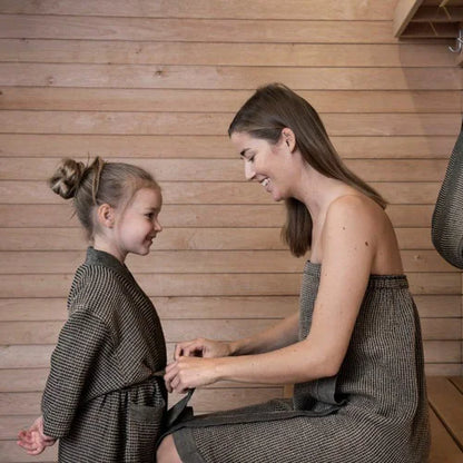 A woman and a young girl wearing matching textured brown and gray patterned dresses or robes, smiling at each other in a warm indoor setting with a wood plank wall background.