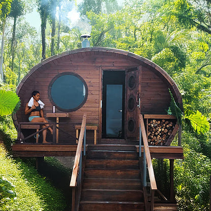 Wooden barrel-shaped cabin in a forest with a person sitting on a bench.