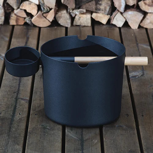 Black pot with wooden handle on a wooden surface with stacked logs in the background
