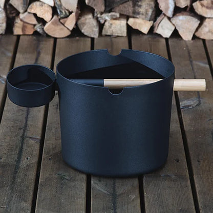 Black pot with wooden handle on a wooden surface with stacked logs in the background