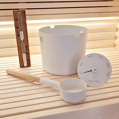 White sauna bucket and ladle with a thermometer on a wooden sauna floor.
