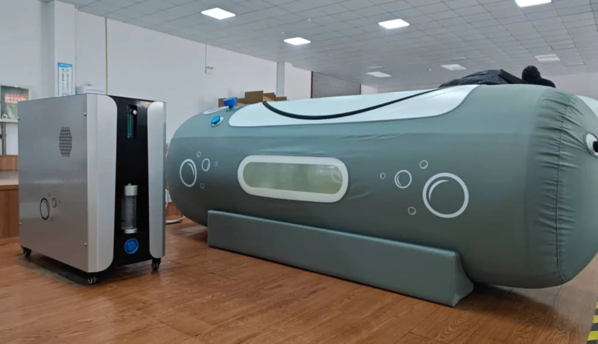 Large inflatable hyperbaric chamber and smaller machine on a wooden floor in a room with tiled ceiling.