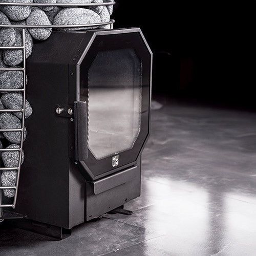 Modern black wood-fire sauna heater insert with glass door in a stone-walled room.