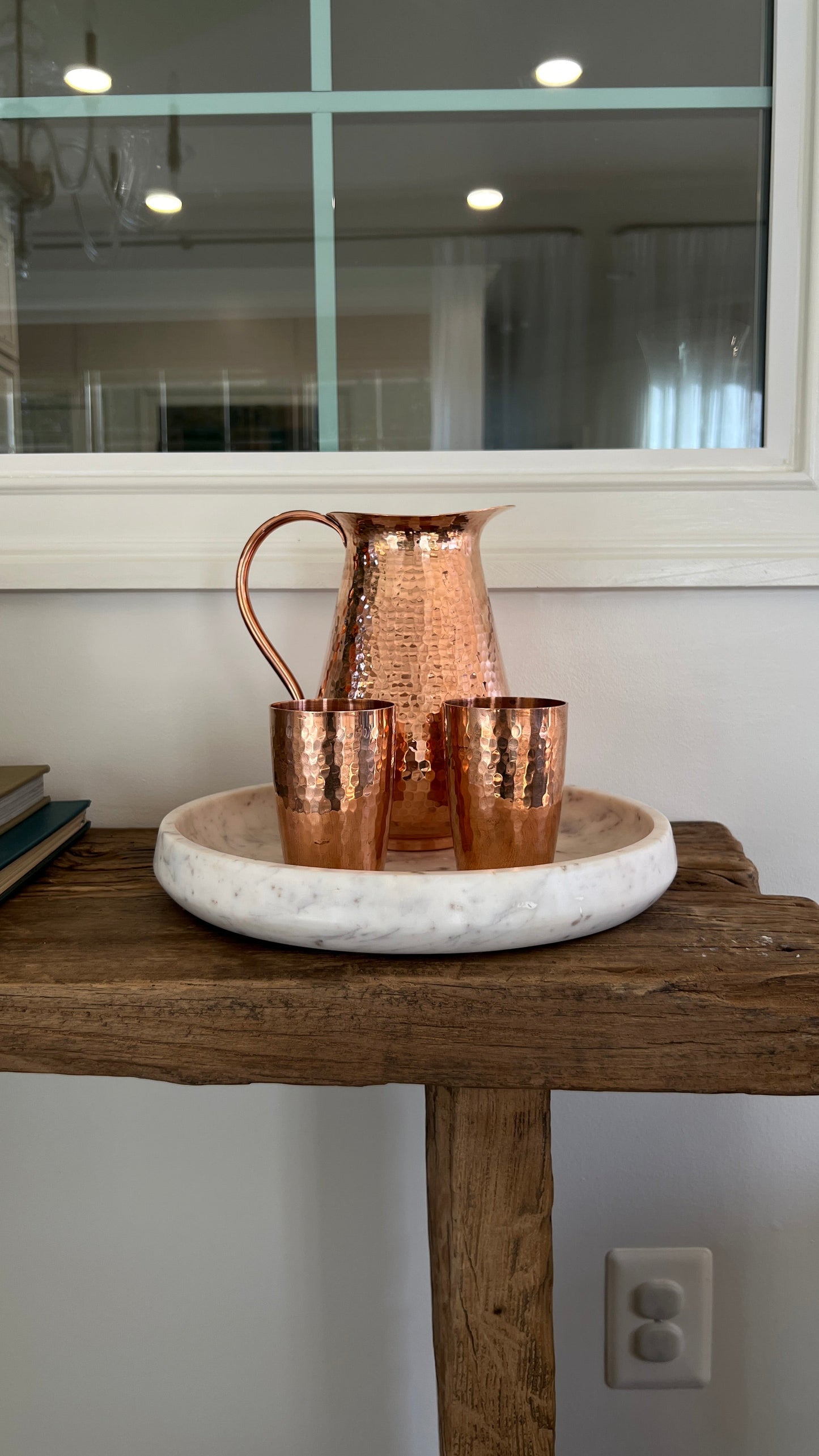 Copper pitcher and cups on a marble tray with a wooden surface and mirror in the background.