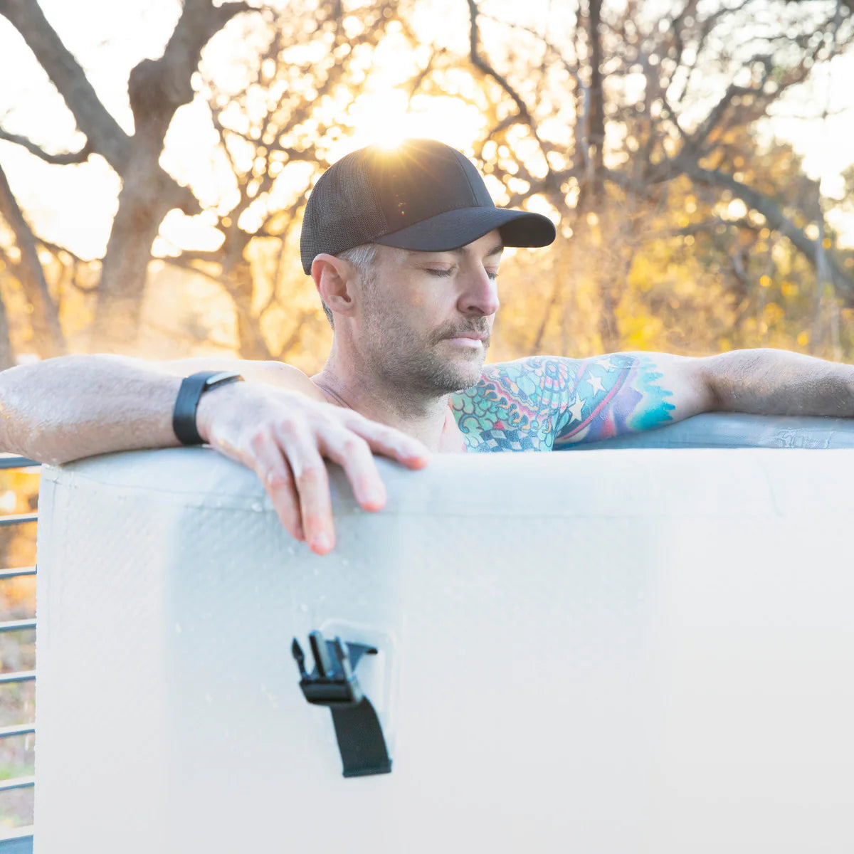 Man with tattoos leaning on a white object outdoors with trees in the background