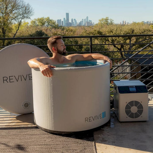 Man relaxing in a Revive portable cold tub on a rooftop with a city skyline in the background.