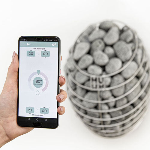 Hand holding a smartphone with a temperature control app next to a sauna heater with stones.
